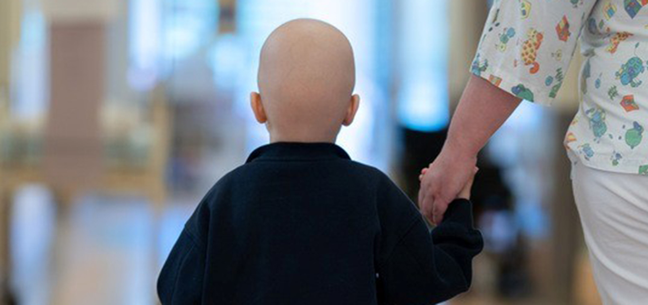 Barn med hårløst hode går hånd i hånd med helsearbeider i lys sykehuskorridor sett bakfra.