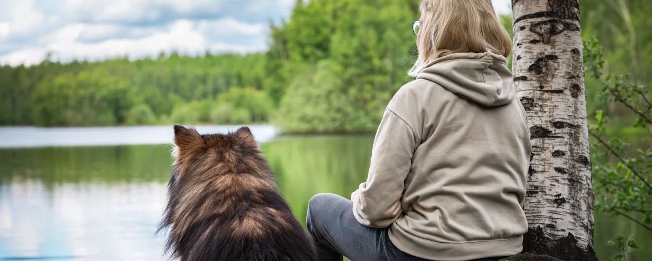 Kvinna sitter med sin hund i en skog och tittar ut över en sjö. Bilden är tagen bakifrån.