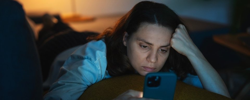 Kvinna ligger på magen i soffan och skrollar i sin mobil på kvällen