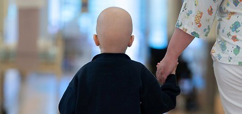 Barn med hårløst hode går hånd i hånd med helsearbeider i lys sykehuskorridor sett bakfra.